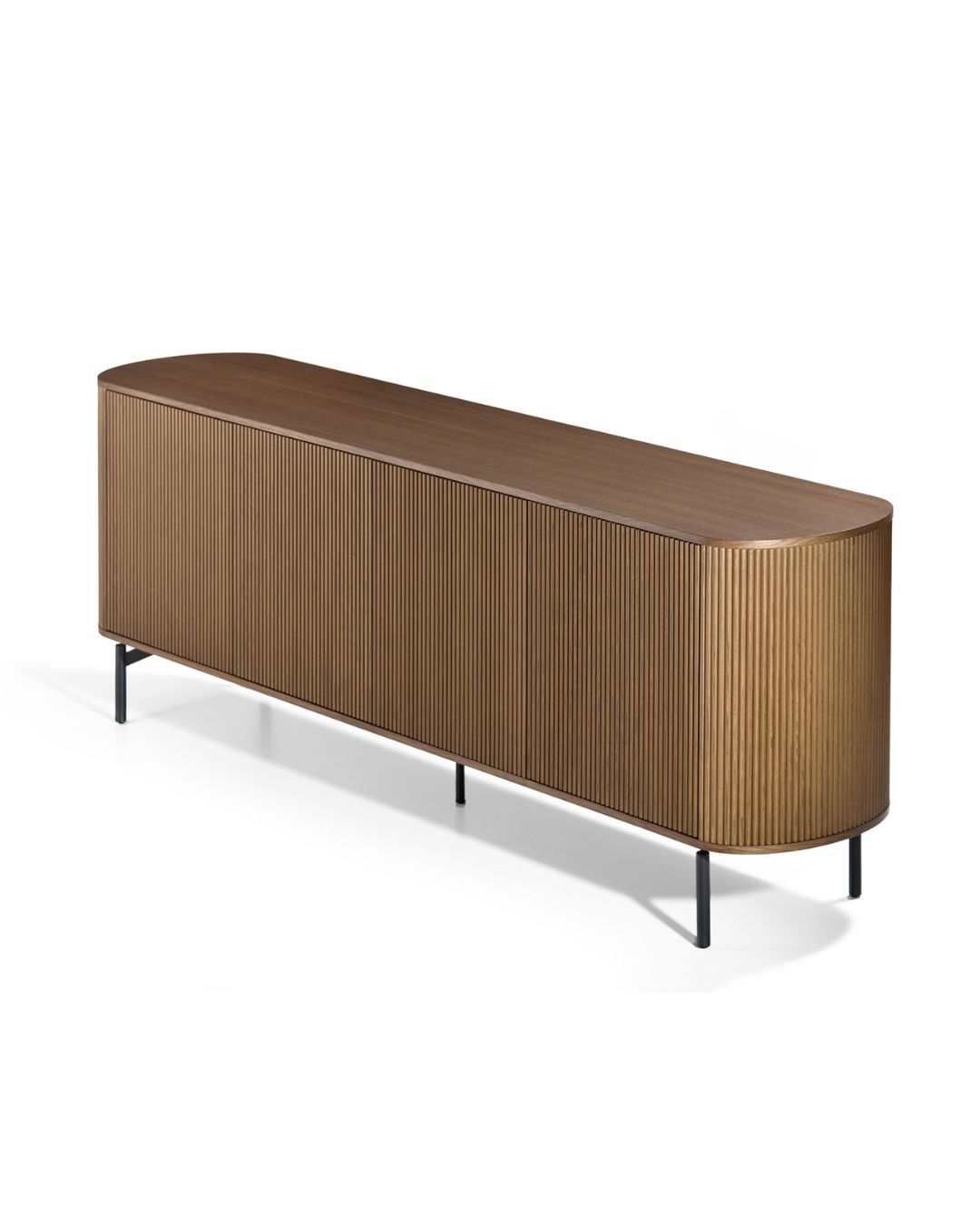 Ilga, minimalistinio dizaino medinė komoda su užapvalintais galais ir vertikaliu frezavimu fasade. Stovi ant plonų juodų metalinių kojelių, paviršius – šiltos riešuto spalvos.